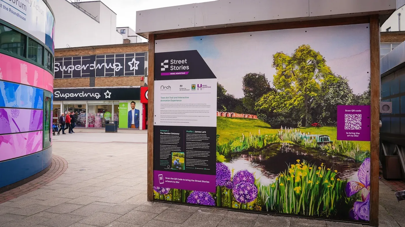 A large printed artwork of a canal-side garden with purple alliums and yellow irises, mounted on a hoarding outside the Roundhouse on Marlowes, with Superdrug behind